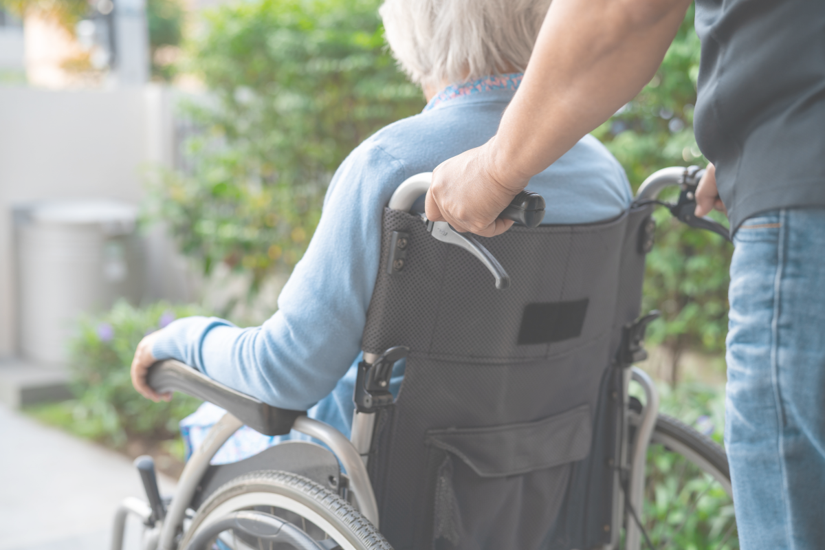 An older adult in a wheelchair being assisted outdoors
